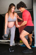 Pregnant woman exercising with dumbbells with personal trainer's help at the gym.
