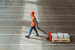 Factory worker, engineer wearing hard hat, vest, work clothes carrying pallet truck with boxes