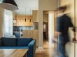 © Svetlana - A blurred man walking in a modern apartment interior with a blue sofa and wooden kitchen cabinets, long exposure