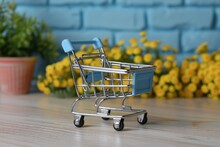 A Small, Metallic Shopping Cart Stands On A Wooden Surface, Its Open Basket Facing Forward, With Vibrant Yellow Flowers In Full Bloom Providing A Cheerful Backdrop To This Charming Setup