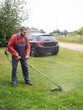 © Александр Лебедько - A man in work overalls mows grass on the lawn with a lawnmower. Taking care of the lawn with a lawnmower in summer on a sunny day. A trimmer cuts the lush green grass on the lawn.