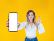 Mobile offer concept, beautiful caucasian blonde curly hair young woman holding showing empty blank screen mobile phone mock up close up to camera. Showing OK sign gesture. Yellow background.