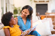 © Monkey Business - Family With Mother And Daughter Sitting On Sofa At Home Reading Book Together