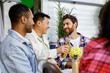 Cheerful multinational company having fun at a picnic. Multisexual friends sitting at outdoor table, celebrating food and drinking drinks. Asian, European and African American mans laughing.