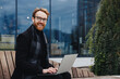 Successful businessman on the background of the business center. Red-haired young guy, in a stylish coat, wireless headphones with a laptop.