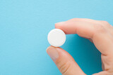 Fototapeta Las - Young adult woman hand fingers holding and showing white pill of c vitamin on light blue table background. Pastel color. Receiving nutrition supplement. Closeup. Top down view.