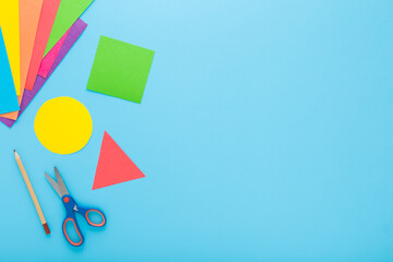 Wall Mural - Colorful geometric shapes, scissors, pencil and application paper on light blue table background. Pastel color. Closeup. Child made different forms for learning. Empty place for text. Top down view.