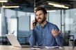 © Tetiana - Smiling young man businessman has a business conversation with clients and partners on the phone, sits in the office at the table and explains actively gesturing with his hands