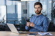 © Tetiana - Portrait of a young serious man working in the office, sitting at a desk, holding a mobile phone and looking confidently into the camera