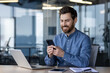 © Tetiana - A smiling young male office worker sits in the office at a desk and uses a mobile phone, writes a message, corresponds