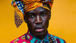 A handsome black man wearing a unique piece of headgear reminiscent of both an African gele and an Asian turban paired with a crisp buttonup shirt and vibrant patterned trousers. His .