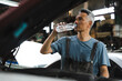 © chokniti - The car mechanic technician is getting exhausted and thirsty, drinking water while checking the engine of car in the factory garage, automobile maintenance during the break time, checking problem fix