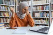 © Liubomir - A young woman in a hijab sits at a library desk, feeling stressed and touching her temples, surrounded by books and a laptop.