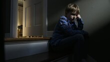 A Sad Young Boy Sits On A Dark Staircase In A Family Home