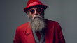 Photo of an old man with a long gray beard and sunglasses wearing a hat, standing fashionably dressed against a gray background, red suit posted
