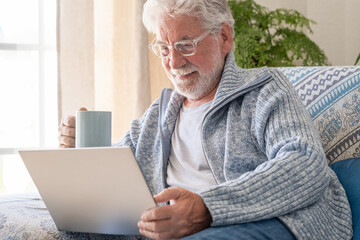 Wall Mural - Senior bearded smiling man sitting on sofa looking at laptop holding a coffee cup - indoor, at home concept - caucasian elderly retired man using technology