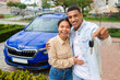 New car. Happy spouses showing automobile key and smiling at camera, hugging standing near auto outdoors. Vehicle dealership