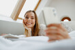 Smiling woman is laying on a bed with a book and a cell phone