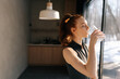 © dikushin - Side view of athletic young woman in sport outfit standing by window and drinking fresh water in morning after fitness training workout exercise at home, enjoying sunny weather and lifestyle.
