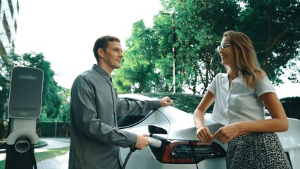 Wall Mural - Young couple travel with EV electric car charging in green sustainable city outdoor garden in summer shows urban sustainability lifestyle by green clean rechargeable energy of electric vehicle innards