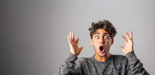 Wall Mural - A man with a shocked expression on his face. He is wearing a gray sweater and has his hands raised in the air. a french young man looking surprised with one of his hands raised