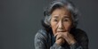 Pensive elderly Asian woman with grey hair and a thoughtful expression, wearing a scarf, embodies grace and wisdom.