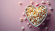 A heart-shaped bowl filled with popped popcorn on a pink background.