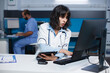 © DC Studio - Female doctor checking patient records on computer while grasping a tablet. In an office, caucasian woman wearing a lab coat is using a digital device and desktop pc to analyze medical data.