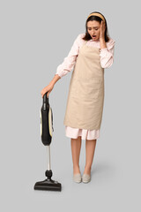 Sticker - Portrait of tired young housewife in apron with vacuum cleaner on grey background