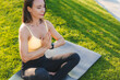 Young caucasian slim woman sitting in lotus yoga pose on fitness mat in sportwear with hands as in prayer in summer park. Calm of healthy young woman breathing