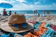A straw hat and sunglasses rest on a colorful towel at a sunny beach with people.