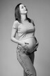 Black and white portrait of young pretty pregnant woman in t-shirt and jeans on gray background.