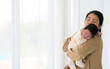 Cute newborn baby girl sleeping on mom shoulder next to white curtain window at home. Safe with mom hug and healthy newborn baby concept