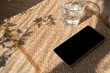 Mobile phone with empty black screen, glass with water on table with sand jute rug background and aesthetic floral sunlight shadows, summer lifestyle home office workspace, copy space