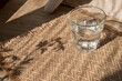 Glass with water on table with sand beige jute rug background with natural floral abstract sunlight shadows, summer lifestyle health care concept, copy space