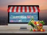 Fototapeta Panele - Full fresh food in shopping basket and supermarket in computer monitor screen with online shopping store graphic and open sign on wooden table over blur of cityscape on warm light sundown