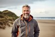 Portrait of a happy man in his 50s wearing a windproof softshell while standing against sandy beach background