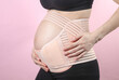 Young Pregnant woman with orthopedic belt for back and belly support. Bandage for pregnant women. Special belt for the 3rd trimester. Studio shot on pink background