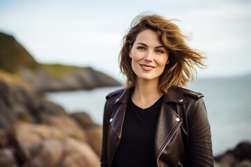 Wall Mural - Portrait of a blissful woman in her 30s sporting a stylish leather blazer while standing against serene seaside background