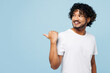 Young cheerful fun happy Indian man he wear white t-shirt casual clothes point thumb finger back aside on area isolated on plain pastel light blue cyan background studio portrait. Lifestyle concept.