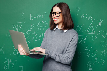 Wall Mural - Young smiling happy smart IT teacher woman wears grey casual shirt glasses use laptop pc computer isolated on green wall chalk blackboard background studio. Education in high school college concept.