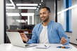 © Tetiana - Young muslim man sitting in office at workplace and doing online yoga, looking at laptop screen and sitting smiling in lotus position