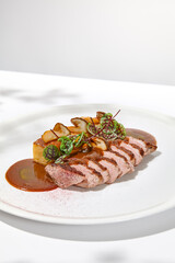 Roasted duck breast with potato gratin and sauce on light background. Fried duck fillet on white plate with hard shadows Elegant summer menu. French cuisine - duck breast with gravy