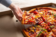 Human caucasian hand takes slice of large pizza from carton box on white wooden table