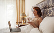 Woman lying in bed with laptop drinking coffee and working. Beautiful blonde student lying on bed in her home and working on her laptop on internet. Girl holding cup of tea with laptop relaxing