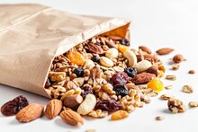Close-up Of A Paper Bag Filled With Assorted Nuts, Dried Fruits, And Granola Bars, Against A White Backdrop.