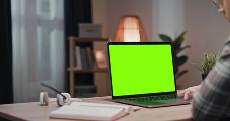 Man using laptop with chroma key screen. Male sitting at table and looking at green mock up of laptop. Man working at modern living room. Male looking at green template laptop display.
