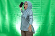 angry young Asian woman wearing hijab and blouse with displeased expression and wanna throw mobile phone over green background
