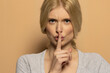 Portrait of beautiful angry woman with long tied blond hair holding index finger on lips on beige background