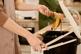 Fototapeta  - Side view of hands of young housewife opening lid of plastic bucket with compost and throwing banana peel after eating fruit